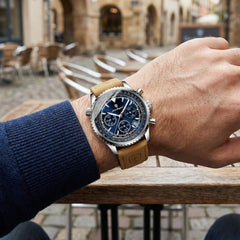 Hand wearing a watch with a brown leather strap on a blurred outdoor background